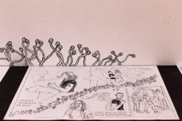 Dolną połowę fotografii zajmuje leżący arkusz z czarno-białym komiksem, ułożony na czarnym tle. Za nim, na białej ścianie, narysowano czarnym pisakiem pnące się kształty przypominające kiełkujące rośliny.