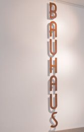 Fotografia przedstawia jedną z prac zakupionych w 2020 roku do Kolekcji Galerii Miejskiej Arsenał. Jest to Bauhaus Rafała Jakubowicza. Praca złożona jest z metalowych pojedynczych liter tworzących w pionie napis: BAUHAUS. Pokryte rdzą litery przymocowane są do białej ściany.